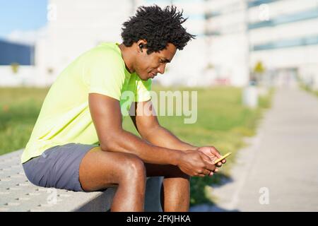 Schwarzer Mann, der sein Smartphone mit einer Übungs-App konsultiert, während er sich von seinem Training ausruhte. Stockfoto