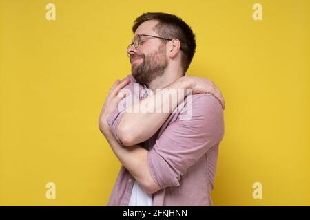 Lustiger bärtiger Mann in runden Gläsern umarmt sich, er leidet unter Narzissmus oder träumt von Liebe, schließt die Augen und lächelt niedlich. Stockfoto