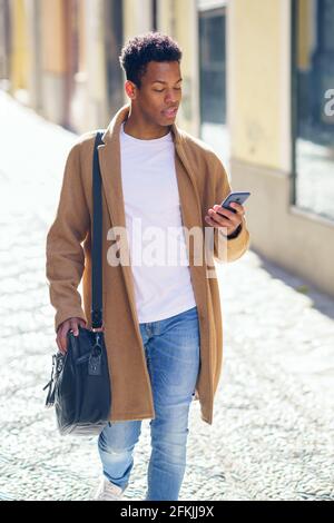 Ein junger schwarzer Mann, der die Straße entlang läuft, trägt eine Aktentasche und ein Smartphone. Stockfoto