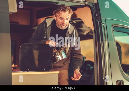 Kaukasische Männer in seinen 50er Jahren mit Wasserkessel im Camper Van, der heißes Wasser für den späten Nachmittag zur Teezeit bereitet. Freizeitfahrzeug Auf Reisen. Stockfoto