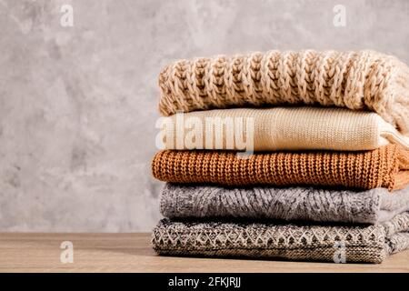 Ein Bund gestrickter Pullover in warmen Pastellfarben mit verschiedenen Strickmustern, die stapelweise auf einem braunen Holztisch gefaltet sind, mit einem grunzten Betonwandhintergrund. Stockfoto