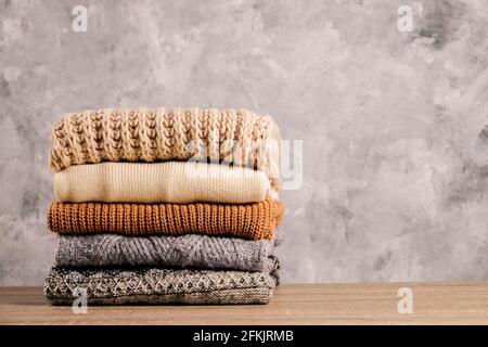 Ein Bund gestrickter Pullover in warmen Pastellfarben mit verschiedenen Strickmustern, die stapelweise auf einem braunen Holztisch gefaltet sind, mit einem grunzten Betonwandhintergrund. Stockfoto