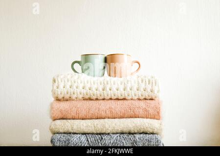 Zwei Tassen Kaffee stehen auf einem Bund gestrickter Pullover in warmen Pastellfarben mit verschiedenen Strickmustern, die im Stapel gefaltet sind. Herbst Winter Strickmode Clother Stockfoto