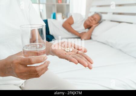 Alte ältere Frau hält Medikamente und Wasser auf dem Bett im Schlafzimmer und ihr Mann schläft daneben. Konzept der Schlafprobleme bei älteren Menschen Stockfoto