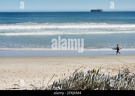 KAPSTADT, Südafrika, 1. Mai 2021: Surfer wandern mit Surfbrett am Strand von Blouberg mit einem Frachtschiff im Hintergrund. Stockfoto