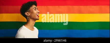 Profilansicht eines schwulen Mannes, der gegen die stolze Flagge steht und lächelt. Banner eines Transgender-Mannes, der Ohrring und Make-up trägt, mit Regenbogenflagge. Stockfoto