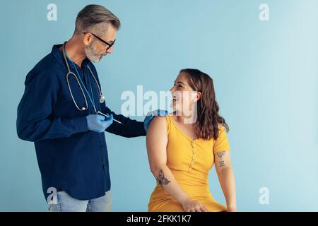 Reifer Arzt hält Injektionsspritze im Gespräch mit Frau Patient auf blauem Hintergrund. Frau erhält Impfstoff von einem männlichen Hausarzt. Stockfoto