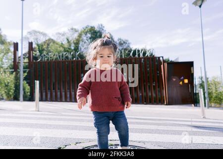 Vorderansicht eines langhaarigen entzückenden Kleinkindes auf einer Blue Jean Hose und einer Purple Jacket, die auf dem Fußgängerüberweg die Straße überquert, allein auf dem Weg zur Schule. Konzept Stockfoto