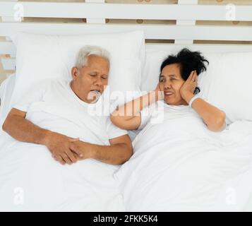 Ältere asiatische Frau, die ihre Augen auflegt und Ohren mit ihren Händen im Bett neben ihrem Mann bedeckt, der schnarcht und Lärm macht, Familienprobleme. Stockfoto