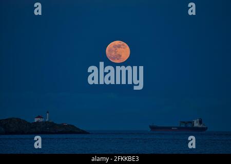 Vollmond über dem Trial Island Lighthouse und Frachtschiff in Twilight-Victoria, British Columbia, Kanada. Stockfoto