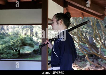 Mann im Samurai-Anzug steht mit Katana auf der Schulter in einem japanischen Gartenhaus. Kirschblütensaison im Botanischen Garten von Moskau Stockfoto