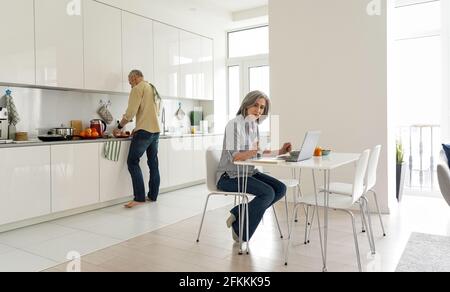 Reife Frau, die von zu Hause aus arbeitet, während ihr Mann in der Küche kocht. Stockfoto
