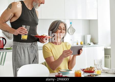 Glückliches älteres reifes Familienpaar, das mit einem Tablet in der Küche frühstückte. Stockfoto