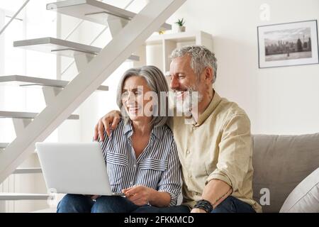 Ein glückliches, älteres Paar im mittleren Alter, das einen Laptop benutzt und zu Hause auf der Couch sitzt. Stockfoto