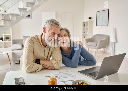 Glückliches reifes älteres Paar lachend, Bonding sitzend am Haupttisch mit Laptop. Stockfoto