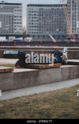 Westminster, London - 2021.05.02: Müde Mann liegt auf der Bank im Uferpark mit seinem Rucksack neben ihm Stockfoto