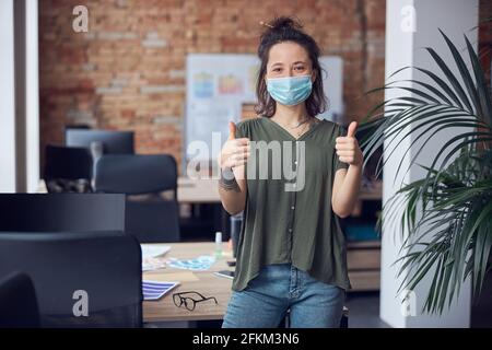 Fröhliche, kreative Frau, die eine Schutzmaske trägt und die Kamera anlächelt und tagsüber im modernen grünen Büro Stumpfe zeigt Stockfoto