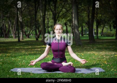 Junge schöne schlanke blonde Frau in lila Gesamt Praxis Yoga Im Sommer grünen Park und zeigt Meditation und Entspannung Asana Sukhasana Stockfoto