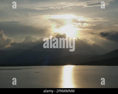 Die gelbe Sonne hinter der Silhouette von Wolke und Berg spiegelt sich bei Sonnenuntergang auf der Seeoberfläche, die Wasseroberfläche funkelt Stockfoto