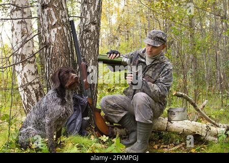 hunter sitzt auf einem umgestürzten Baum und gießt Tee aus einer Thermoskanne, die neben seinem Hund sitzt Stockfoto