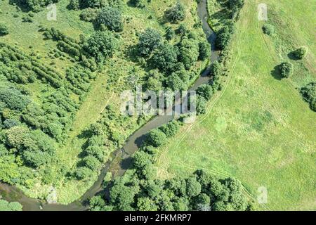 Kleiner Fluss, der sich durch grüne Felder schlängelt, umgeben von Mischwäldern. Luftaufnahme an sonnigen Tagen. Stockfoto