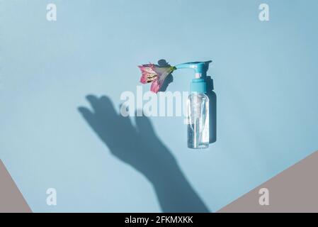 Spender mit transparentem Gel und Blume auf hellblauem Hintergrund. Schatten Silhouette einer weiblichen Hand. Stockfoto