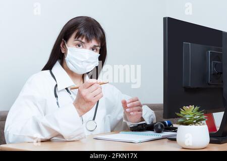 Eine Ärztin, die eine medizinische Schutzmaske trägt, schaut Sie an, während sie an einem Tisch im Büro sitzt. Der Arzt hört auf die Symptome des Patienten Stockfoto