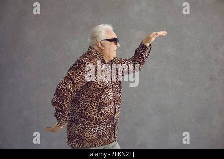 Seitenansicht eines lustigen älteren Mannes im Leopardenhemd, der tanzt Im Studio mit grauem Hintergrund Stockfoto