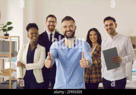 Lächelnder Anführer, der mit Daumen nach oben steht und mit einem vielfältigen Geschäftsteam zusammensteht Im Büro Stockfoto