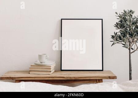 Vertikaler schwarzer Bilderrahmen-Nachbau auf Vintage-Bank, Tisch. Tasse Kaffee auf Bücherstapel. Eingetopfter Olivenbaum. Weißer Wandhintergrund. Skandinavisch Stockfoto