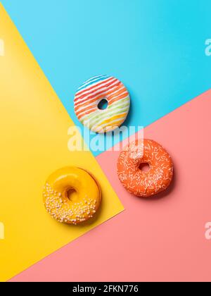 Kreatives Layout aus köstlichen glasierten Donuts. Vertikale flache Lay - Donuts oder Donuts auf buntem Hintergrund mit Kopierraum. Blauer, gelber und rosafarbener geometrischer Hintergrund. Stockfoto