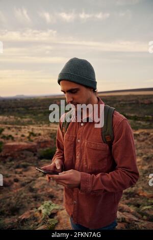 Hübscher junger Mann, der Smartphone mit Kappe und Rucksack hält Stockfoto