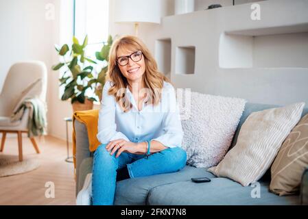 Aufnahme einer glücklichen, reifen Frau, die weißes Hemd und Jeans trägt, während sie sich zu Hause auf dem Sofa entspannt. Stockfoto