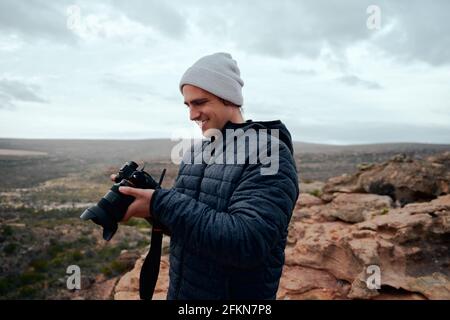 Männlicher Reisefotograf überprüft Fotos nach der Aufnahme im Stehen auf der Kamera Auf dem Berg mit Mütze Stockfoto