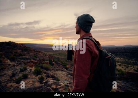Junger männlicher Wanderer, der eine Wasserflasche mit Rucksack und Kappe hält Blick auf den Sonnenaufgang am Morgen Stockfoto