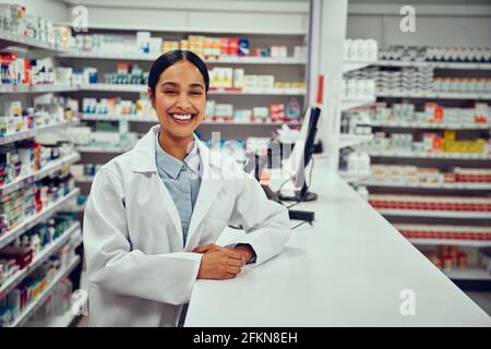 Porträt einer lächelnden, glücklichen, selbstbewussten jungen Apothekerin, die sich darauf stützt Ein Schreibtisch in der Apotheke Stockfoto
