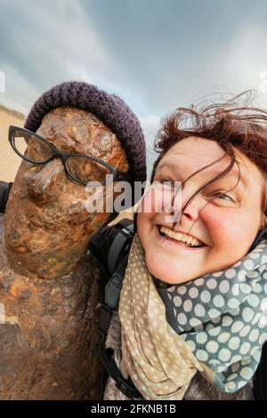 Frau grinst neben der gusseisernen Skulptur eines Mannes, der einen wolligen Hut und eine Brille trägt. Ein anderer Ort ist Antony Gormley am Crosby Beach Stockfoto