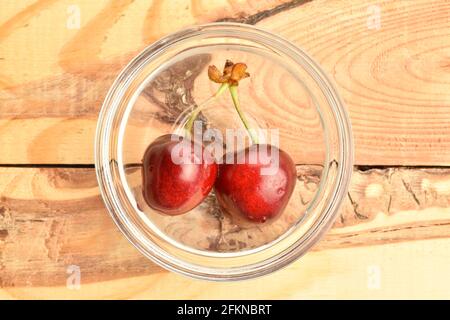 Zwei dunkelrote, süße, saftige Kirschen in einer Glasschale auf einem Holztisch, Nahaufnahme, Draufsicht. Stockfoto