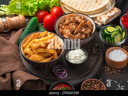 Pommes frites, gebratenes Fleisch, Pita-Brot, Gemüse und Gewürze auf dunklem Hintergrund. Eine vollständige Mahlzeit oder Zutaten für Shawarma, Burrito, Gyros. Nahaufnahme. Stockfoto