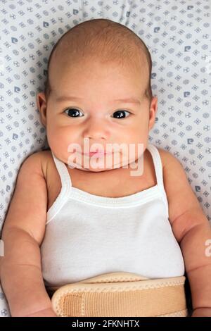 Baby in einer Nabelbruch-Bandage. Süßer kahler Säugling auf dem Bett Stockfoto