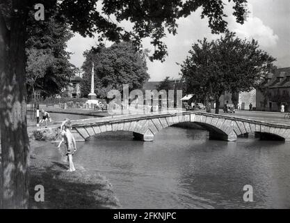 Die 1950er Jahre, ein historisches Bild aus dieser Zeit, sehen wir kleine Kinder, die am Ufer des sanft fließenden Flusses Windrush in Bourton-on-the-Water spielen, einem hübschen Dorf in den englischen Cotswolds. Bekannt für seine niedrigen Brücken und traditionellen Steinhäuser, ist das Dorf ein charmanter Ort zum Besuchen und wird oft als das "Venedig der Cotswolds" bezeichnet. Stockfoto