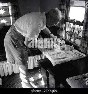 In den 1950er Jahren, historisch, in einem engen Raum, vielleicht in einem kleinen Ferienhaus oder einer alten Schäferhütte, ein Herr, der ein leichtes Essen zubereitet, indem er ein Stück Huhn mit dem Licht aus einem kleinen Schiebefenster, mit der unteren Hälfte mit Buntglasfenster, Stockfoto