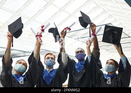 Diverse Gruppe von Hochschulabsolventen, die Hüte in die Luft werfen und Masken während der Abschlusszeremonie in Innenräumen tragen, Platz kopieren Stockfoto