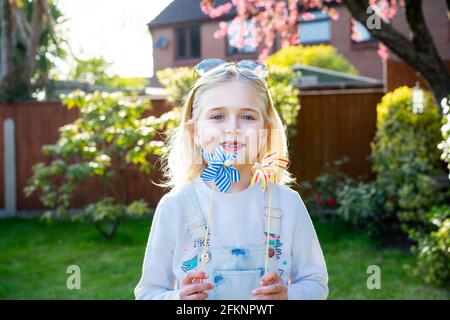 Lächelndes zahnloses kleines Mädchen mit bunten Spielzeug Windmühlen viel Spaß im Garten. Aktivitäten im Freien. Frohe Kindheit. Selektiver Fokus, Kopierbereich Stockfoto