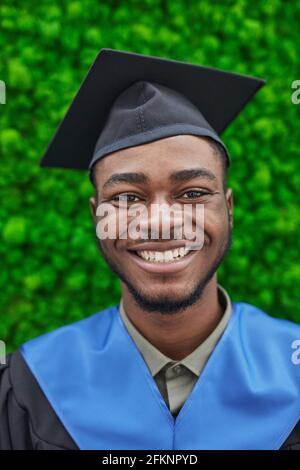 Vertikales Nahaufnahme-Porträt eines glücklichen afroamerikanischen Mannes mit Graduierung Mütze und lächelnd auf die Kamera, während sie gegen grüne Pflanze steht Hintergrund Stockfoto