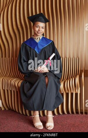 Vertikale Ganzkörperportrait mit lächelnder junger Frau mit Graduierung Beim Posen im Innenbereich mit einem Lächeln auf die Kamera Stockfoto