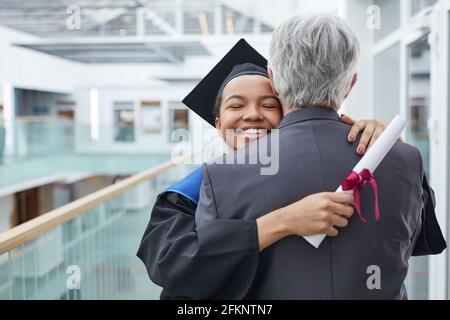 Porträt einer glücklichen afroamerikanischen Frau im Abschlusskleid, die Vater oder College-Professor in Innenräumen umarmt, Kopierraum Stockfoto