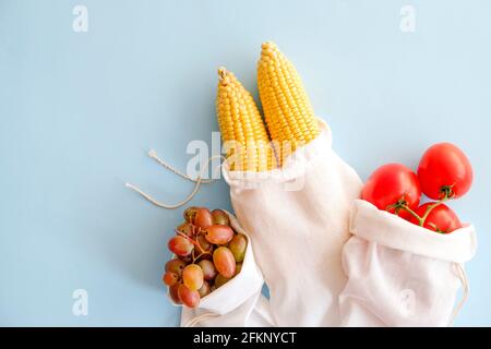 Zwei Maiskolben, Trauben und Zweig von Kirschtomaten in drei wiederverwendbaren umweltfreundlichen Baumwollbeuteln mit Krawatten auf hellblauem Hintergrund. Keine Verschwendung c Stockfoto
