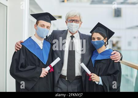 Waist-up-Porträt von zwei Hochschulabsolventen, die Masken tragen, während Posiert mit reifen Professor in Innenräumen Stockfoto