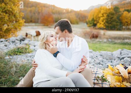 Das Paar küsst sich, während es auf einer Decke vor dem Hintergrund sitzt Von Pferden, die im Herbstwald grasen Stockfoto